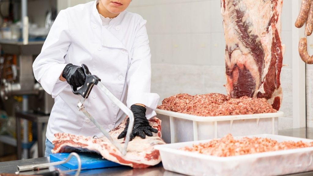 A butcher cutting a large piece of meat on a cutting board using a butcher saw
